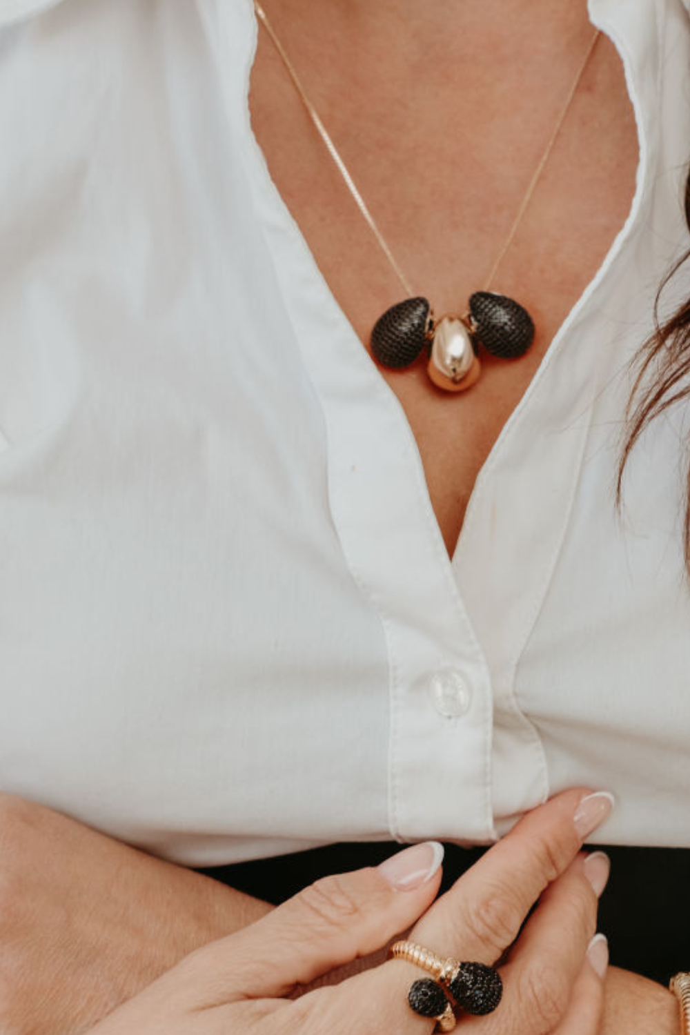 A close up lifestyle shot of a woman wearing a gold necklace with three textured black zirconia and metallic gold beads, paired with a white button down shirt.